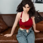 Amalia Sex Doll 166 cm IronTech, assise sur canapé, cheveux bruns bouclés, top rouge, jean décontracté.