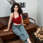 Amalia Sex Doll 166 cm IronTech, assise sur canapé, cheveux bruns bouclés, top rouge, jeans décontracté.