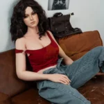 Amalia Sex Doll 166 cm en pose assise, cheveux bruns bouclés, top rouge, jeans décontracté.