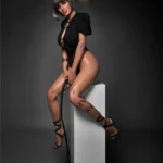 Amova Sex Doll 165 cm IronTech en tenue noire, assise sur un cube, cheveux courts, tatouage floral.