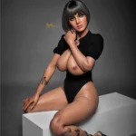 Amova Sex Doll 165 cm IronTech Doll en body noir, assise sur un cube blanc, cheveux courts.