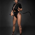 Amova Sex Doll 165 cm IronTech en pose debout, tenue noire, lunettes, fouet en main.