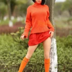 Aurabella Sex Doll 167 cm en jupe rouge et pull orange, posant debout près d'un arbre.
