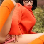 Aurabella Sex Doll 167 cm IronTech, pose assise, pull orange, bas orange, lunettes noires.
