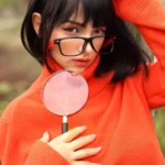 Aurabella Sex Doll 167 cm IronTech en pull orange, lunettes noires, tenant une loupe.