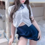 Bellora Sex Doll 168 cm IronTech Doll en uniforme scolaire, posée sur un lit, portant des lunettes.