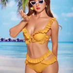 Elodya Sex Doll 168 cm IronTech en bikini jaune, lunettes de soleil, plage en arrière-plan.