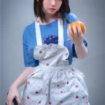 Elyzabelle Sex Doll 164 cm en tablier à cœurs, tenant une orange, pose debout, regard vers l'avant.