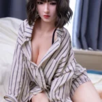 Lilina Sex Doll 165 cm IronTech Doll assise sur un lit, chemise rayée ouverte, cheveux courts.