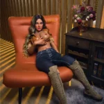 Luméa Sex Doll 165 cm IronTech Doll, assise sur fauteuil orange, chemise ouverte, jean et bottes.