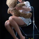 Lumiria Sex Doll 165 cm assise sur une chaise, tenant un ours en peluche, cheveux courts gris.