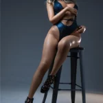 Real Doll ultra réaliste en silicone Lunora, 166 cm, par IronTech Doll. Élégamment assise sur un tabouret, portant un maillot de bain bleu écailles et des talons noirs.
