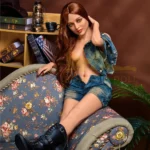 Luxima Sex Doll 150 cm IronTech, assise sur canapé, veste en jean, pose détendue.