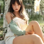 Mabilie Sex Doll 167 cm posée assise contre un arbre, robe blanche, ambiance extérieure naturelle.