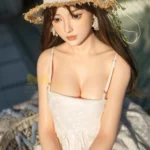 Ninabelle Sex Doll 166 cm en robe blanche, chapeau de paille, assise en extérieur.