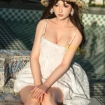 Ninabelle Sex Doll 166 cm assise en robe blanche, chapeau de paille, jardin ensoleillé.