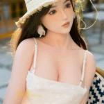 Ninabelle Sex Doll 166 cm IronTech en robe blanche, chapeau de paille, assise en extérieur.