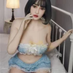 Alia Sex Doll 159 cm IronTech Doll TPE assise sur un lit, en lingerie bleue et blanche.