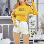 Alicine Sex Doll 163 cm IronTech Doll TPE en pull jaune et short blanc dans une chambre moderne.