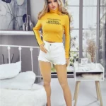 Alicine Sex Doll 163 cm IronTech, debout en short blanc et pull jaune, intérieur moderne.