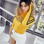 Alicine Sex Doll 163 cm IronTech Doll TPE debout, main dans les cheveux, portant un haut jaune.