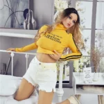 Alicine Sex Doll 163 cm IronTech TPE posée sur un lit, vêtue d'un haut jaune et short blanc.