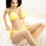 Alya Real Doll 169 cm IronTech TPE assise en bikini jaune sur plancher clair, cheveux noirs bouclés.
