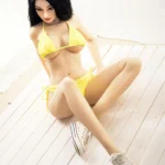 Alya Real Doll 169 cm IronTech en bikini jaune assise sur le sol, cheveux noirs ondulés.