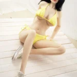 Alya Real Doll 169 cm IronTech en bikini jaune, posée assise sur un sol en bois.