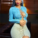 Alyona Sex Doll 164 cm en jupe beige et top bleu, assise sur canapé, regard intense.