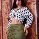 Amelia Sex Doll 160 cm IronTech Doll TPE en jupe verte et chemisier à pois, posée contre un mur.