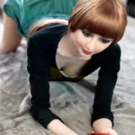 Aurélie Real Doll 165 cm IronTech en position à genoux, robe verte, regardant un smartphone.