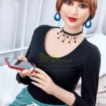 Aurélie Real Doll 165 cm assise, tenue décontractée, tenant un smartphone, boucles d'oreilles bleues.