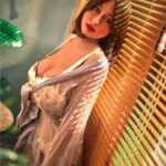 Eliya Sex Doll 163 cm en robe transparente, appuyée contre des stores en bois, lumière douce.