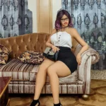 Eloda Sex Doll 156 cm IronTech Doll TPE assise sur canapé, robe noire, lunettes.