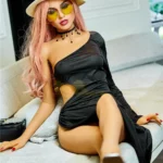 Elodora Sex Doll 165 cm IronTech en robe noire, chapeau blanc, lunettes jaunes, posée sur canapé.