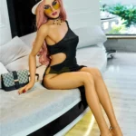 Elodora Sex Doll 165 cm IronTech en robe noire, assise sur canapé, chapeau blanc.