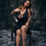 Elody Sex Doll 158 cm IronTech, assise sur chaise, robe noire, cheveux longs bruns.