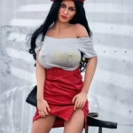 Enora Sex Doll 163 cm en jupe rouge, t-shirt gris, béret rouge, posée sur chaise transparente.