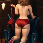 Evabella Sex Doll 158 cm en lingerie rouge, agenouillée sur un fauteuil, dos visible.