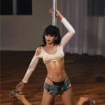 Florine Sex Doll 168 cm IronTech posée à genoux, tenue sexy, près d'une barre de pole dance.