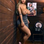 Lilaya Sex Doll 158 cm IronTech Doll TPE debout contre un mur en bois, robe grise, talons roses.