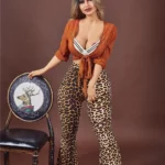 Lovena Sex Doll 156 cm IronTech Doll TPE debout, chemise orange, pantalon léopard, main sur chaise.