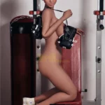 Lulabelle IronTech Doll 163 cm posée nue sur un banc de musculation, appareil photo vintage.