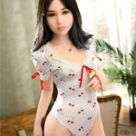 Luludoll Sex Doll 165 cm en body blanc à motifs cerises, debout près d'une chaise.