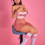Lumabelle Sex Doll 153 cm IronTech assise sur tabouret, jupe rose, écouteurs roses.