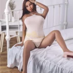 Lumizelle Sex Doll 163 cm posée sur lit, robe blanche, jambes croisées, ambiance chambre.