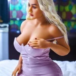 Nimaya Sex Doll 156 cm IronTech en robe violette, assise sur un lit, cheveux blonds ondulés.