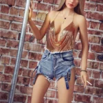 Nimya Sex Doll 163 cm IronTech, pose debout, top doré, short en jean, fond mur de briques.