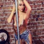 Nimya Sex Doll 163 cm IronTech en tenue glamour, appuyée contre un poteau, fond de briques.